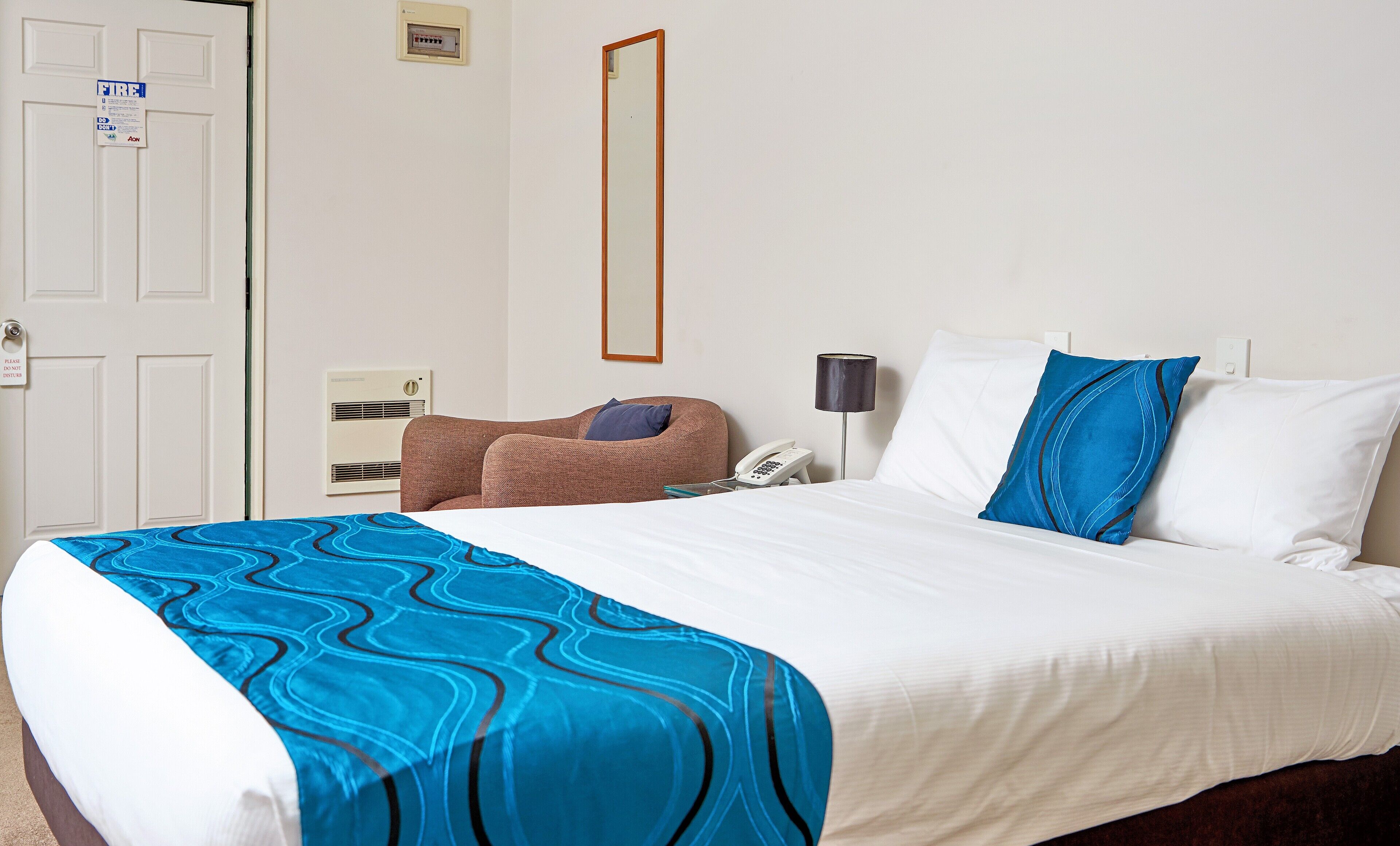 A hotel room with a bed, a chair, a bedside table, a lamp, and a wall-mounted phone.