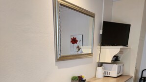 Una habitación pequeña y limpia con una mesa de madera y una silla negra, un escritorio blanco con una computadora y una planta pequeña, un cuadro en la pared y un televisor colocado sobre el escritorio.