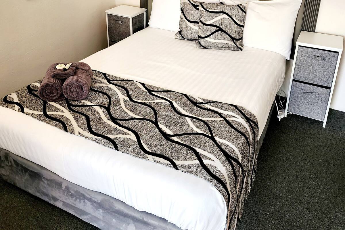 A neatly made bed with a patterned bedspread, two pillows, and a decorative item on the bed. There are two bedside tables with lamps and a small drawer unit beside the bed.