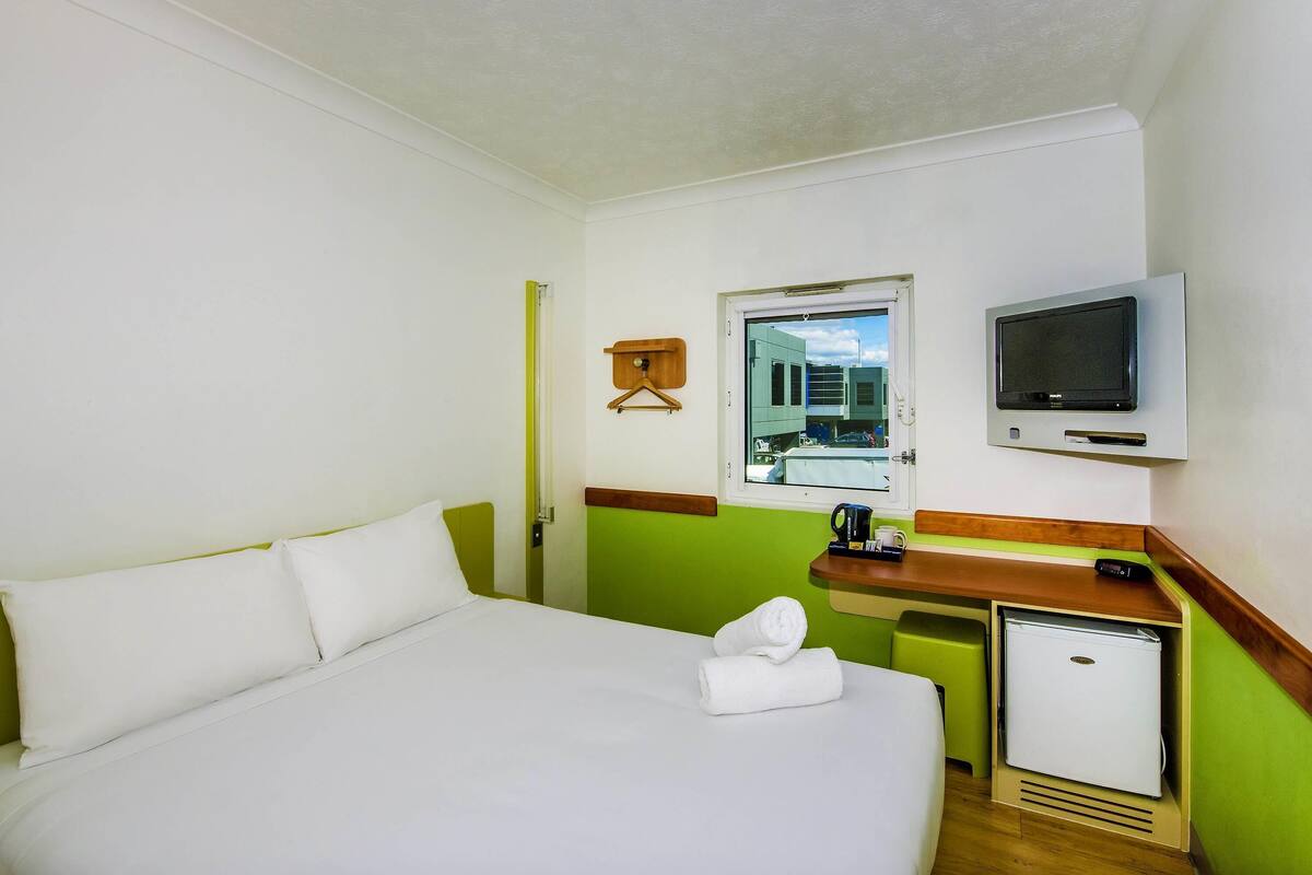 Une chambre d’hôtel équipée d’un lit, d’une télévision, d’un petit réfrigérateur et d’une fenêtre donnant sur des immeubles.