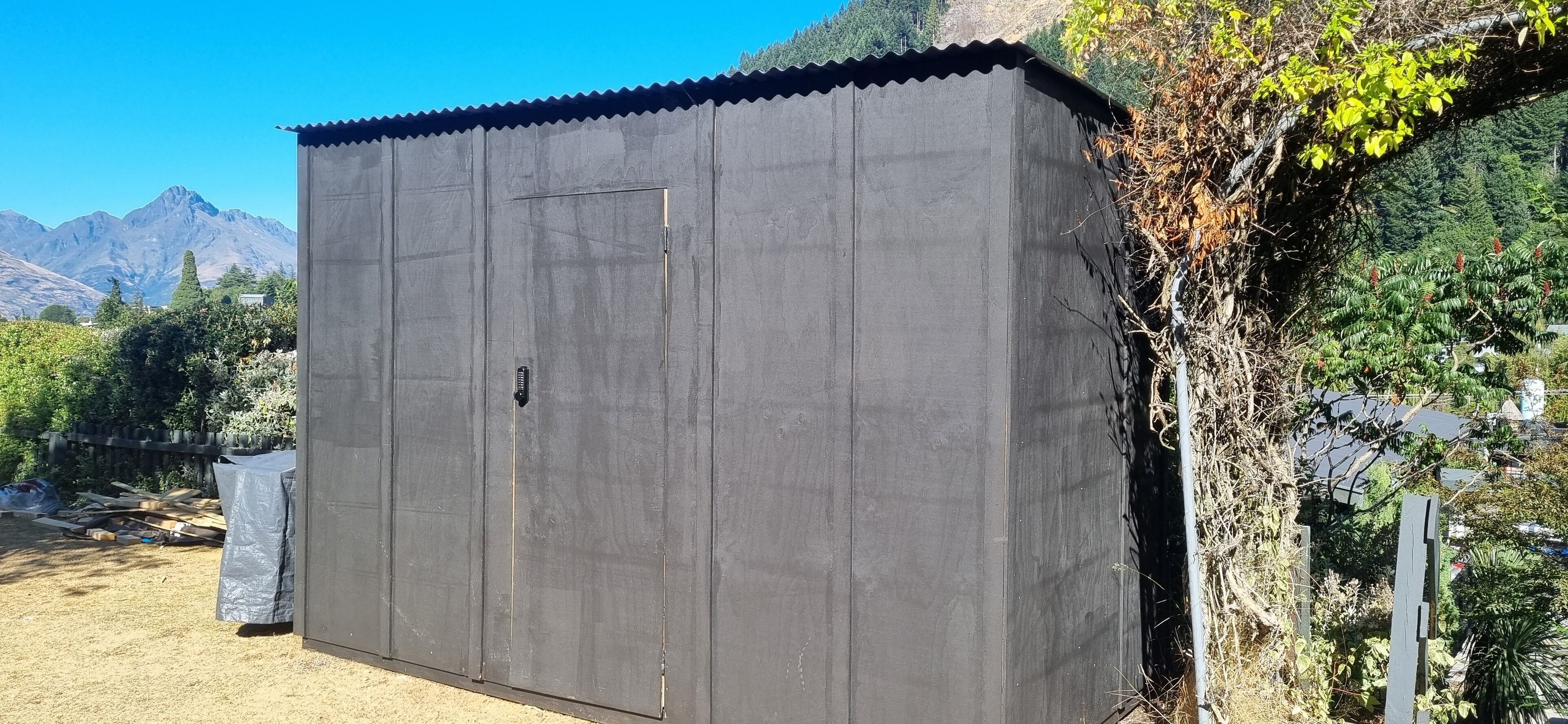 A large, dark-colored metal shed with a corrugated roof, situated in a garden with mountains in the background.