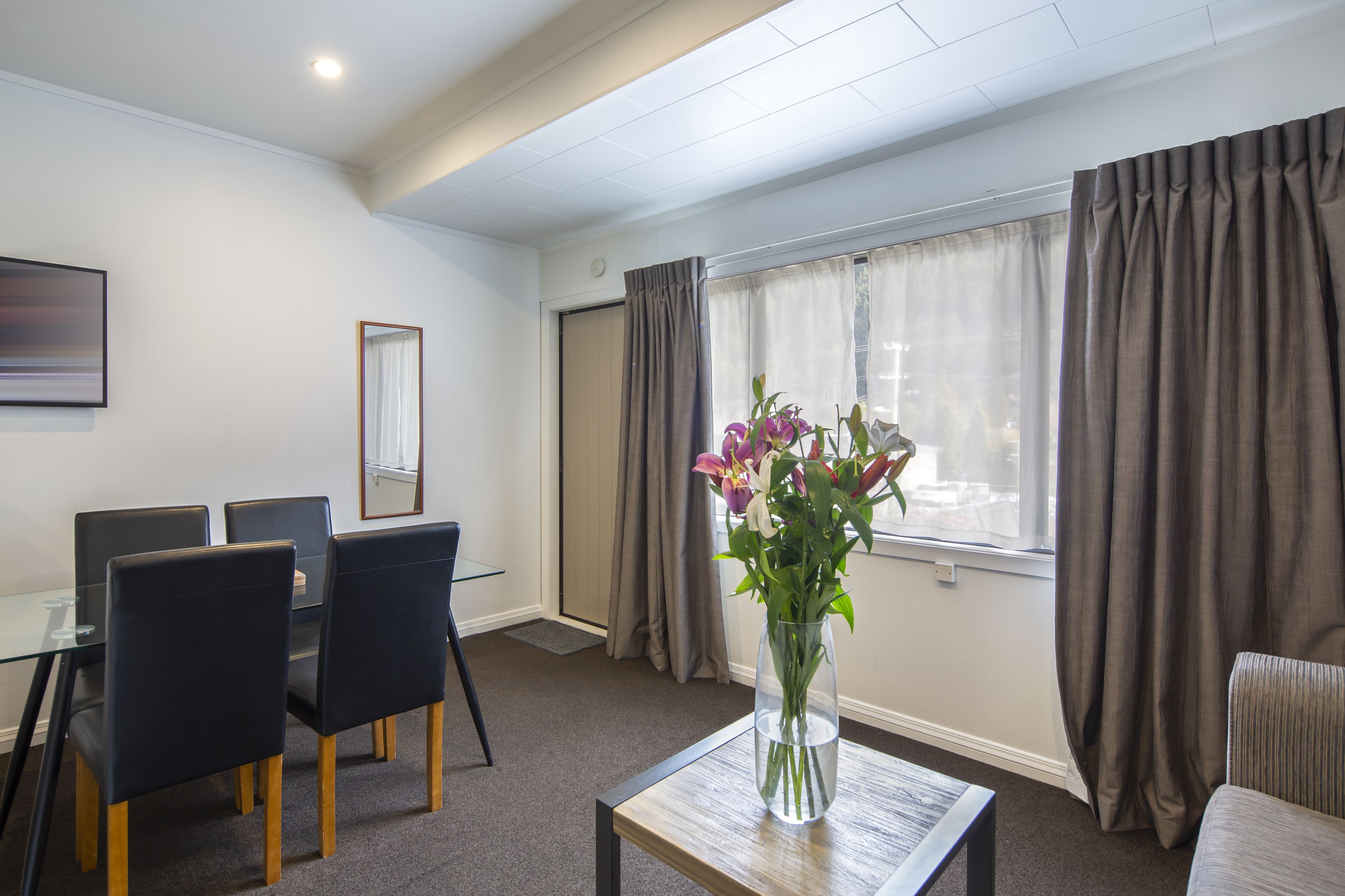 Habitación de hotel con una mesa de comedor, sillas, un jarrón con flores, una ventana con cortinas y vistas a la ciudad.