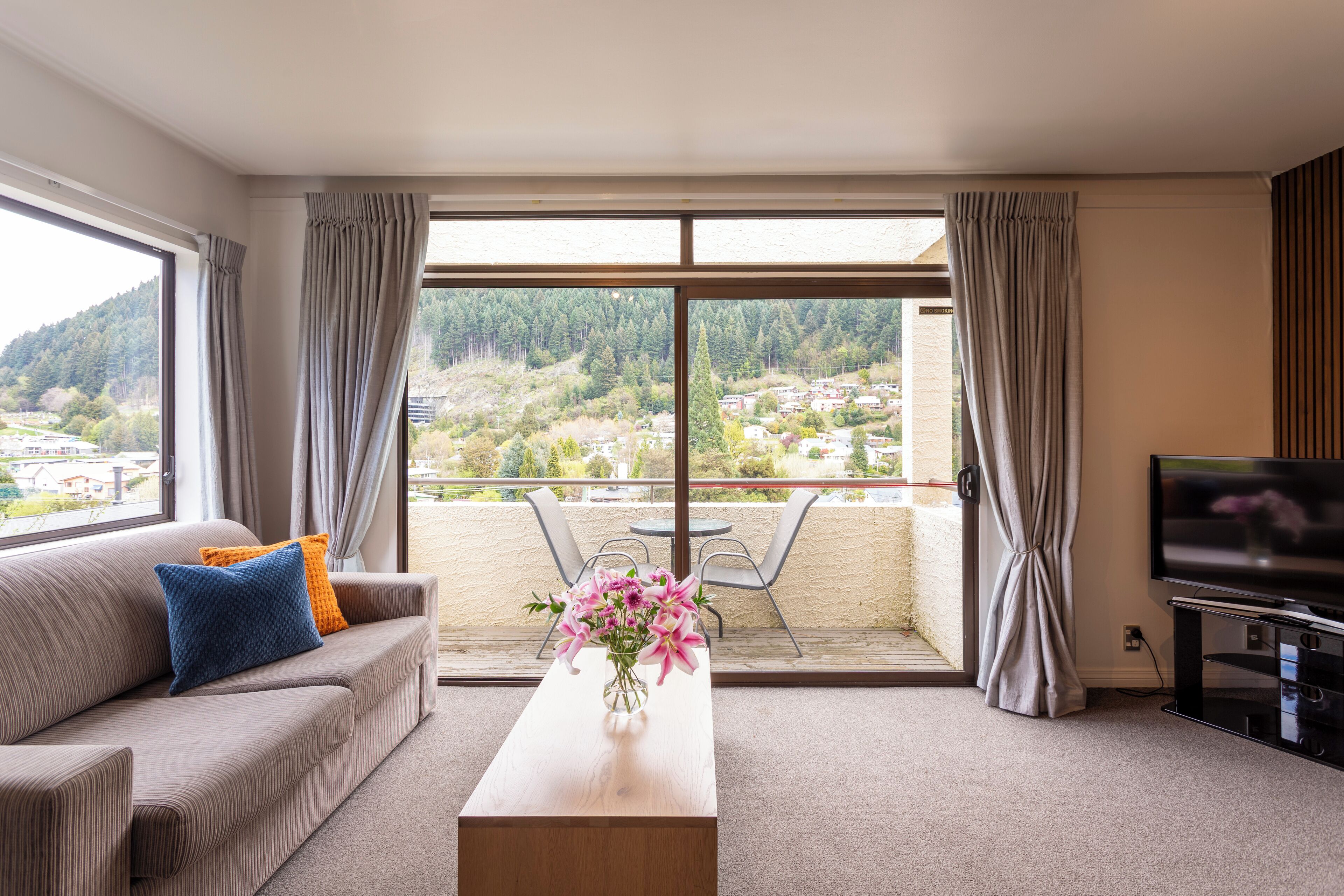 Un salon moderne avec un canapé, une table basse ornée d’un vase de fleurs, un téléviseur et un balcon offrant une vue sur la ville et les collines.