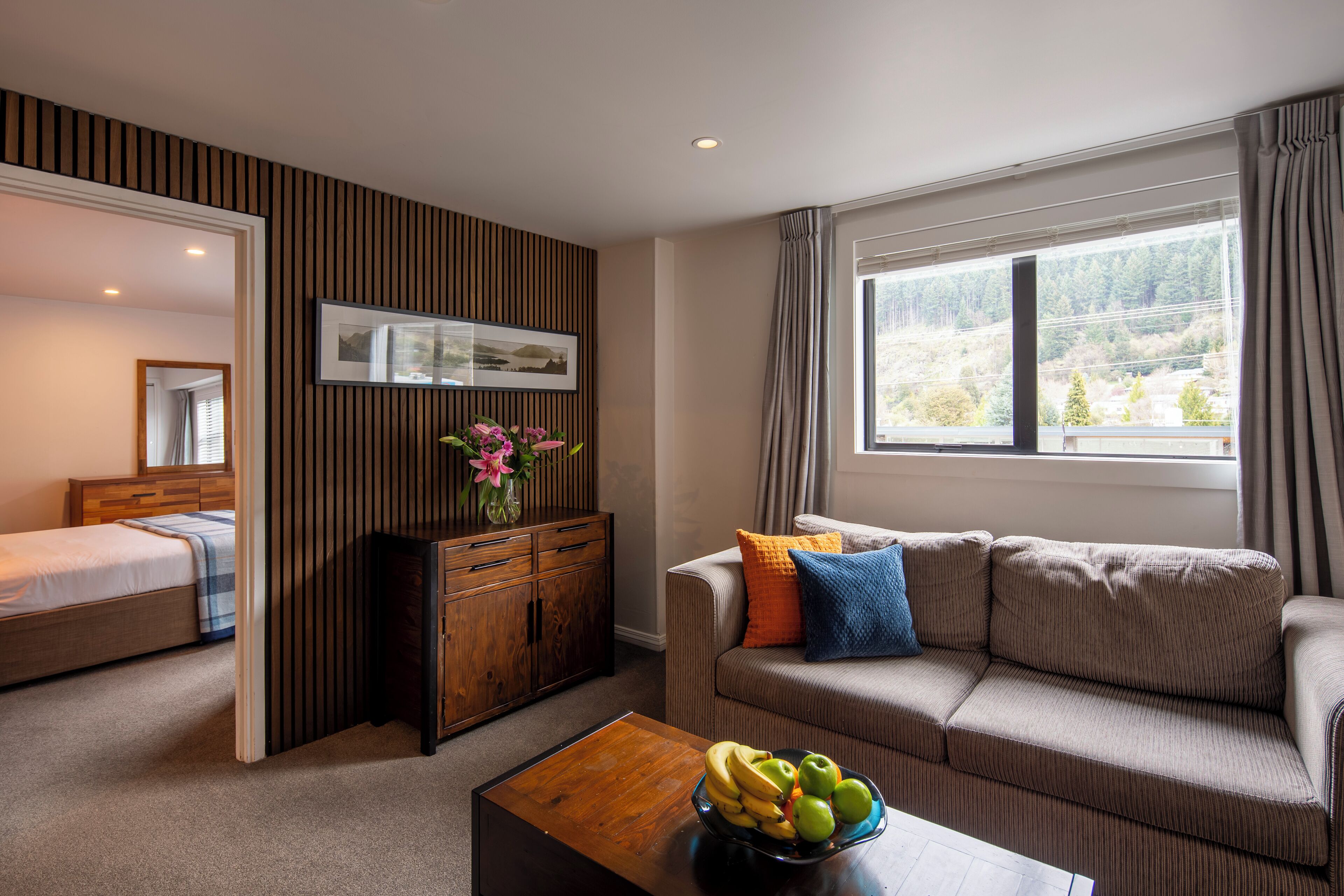 Une chambre d’hôtel moderne comprenant un lit, un canapé, une table basse en bois avec des fruits, une commode et une grande fenêtre donnant sur des arbres.