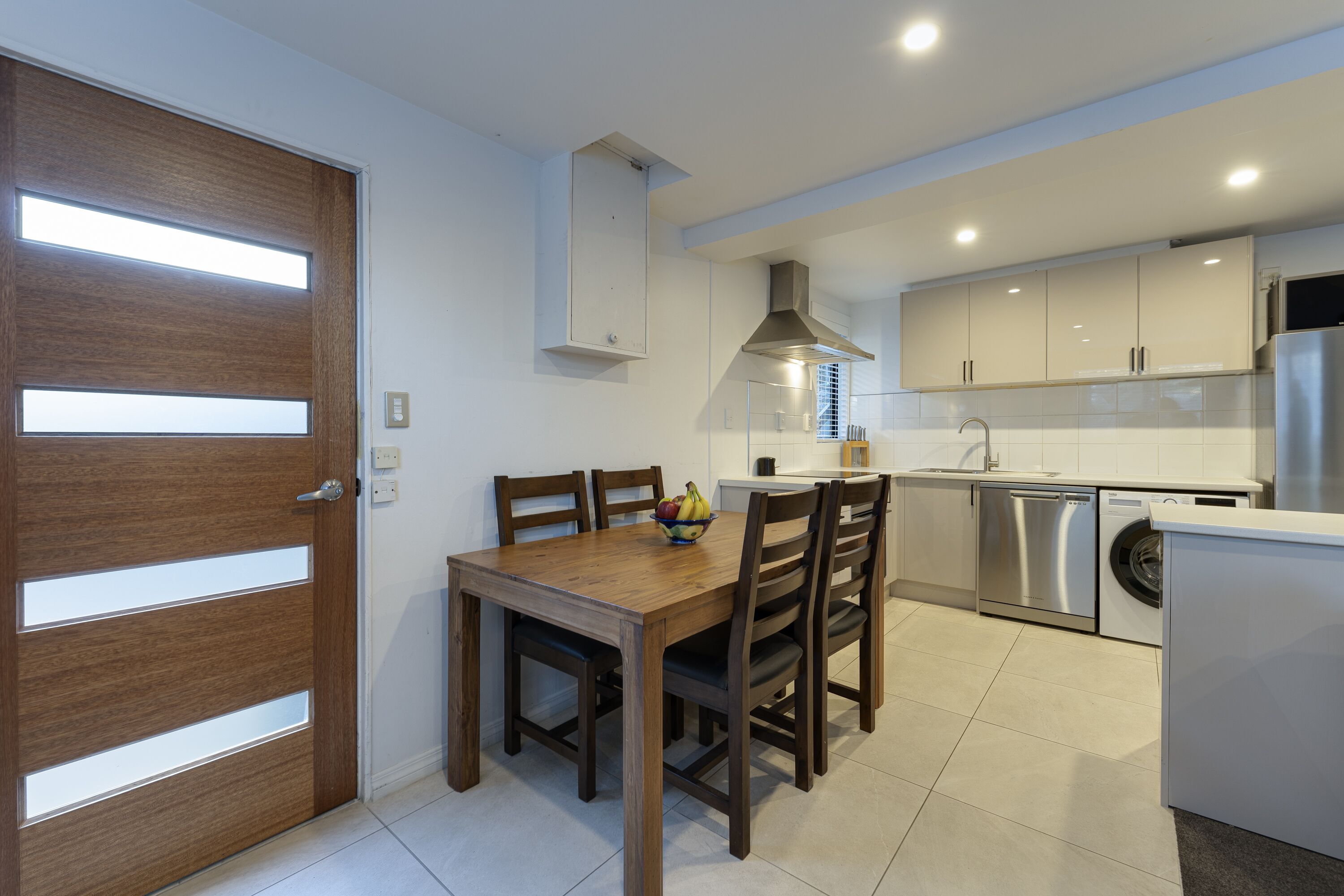 Una cocina moderna con una mesa de comedor de madera, sillas, un frutero, electrodomésticos de acero inoxidable y piso de baldosas blancas.