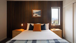 A neatly arranged bedroom with a large bed, two bedside tables, a nightstand, a framed picture on the wall, and a window providing a view. - Abba Court Motel & Apartments (Queenstown)