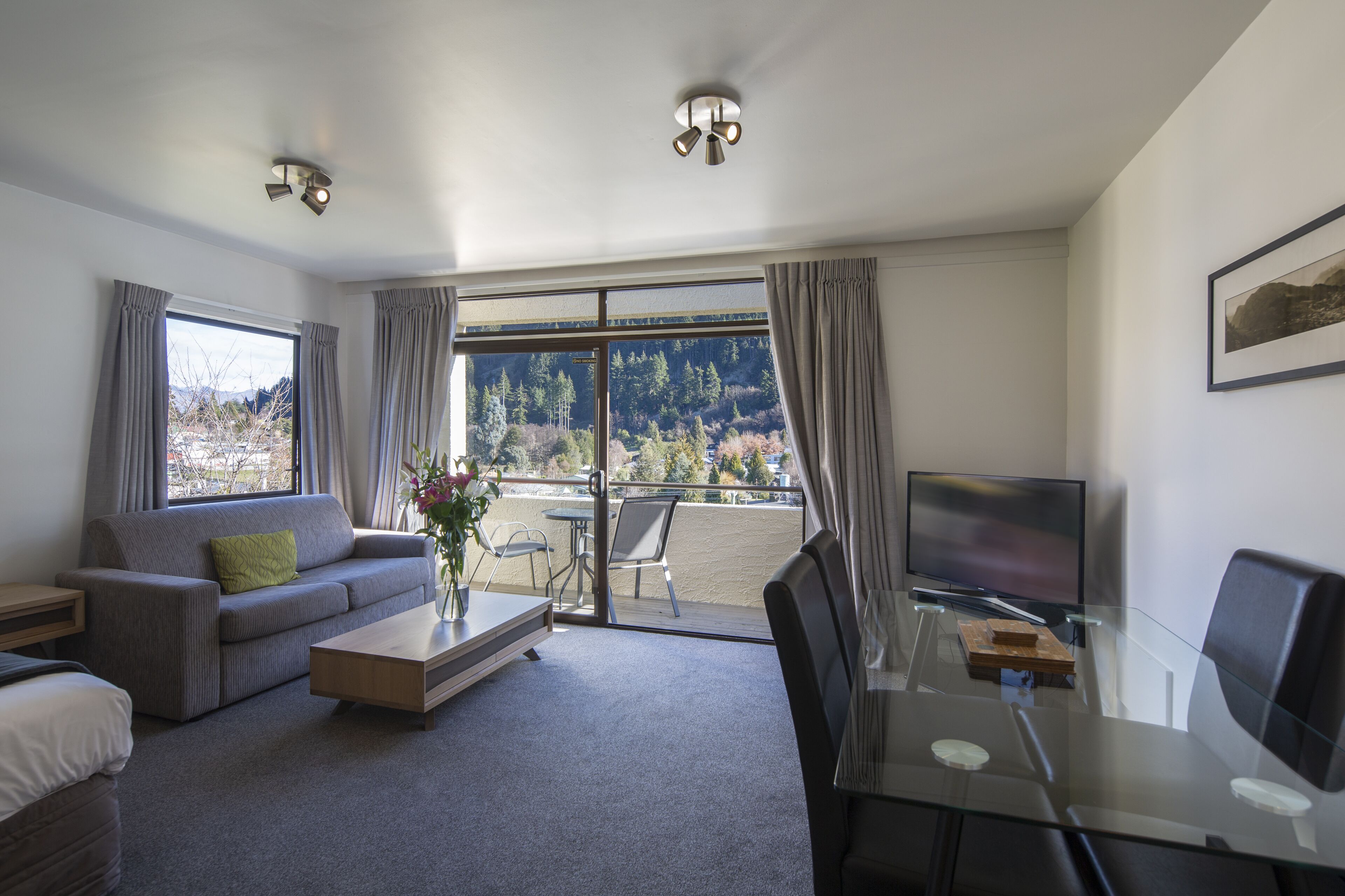 Ein modernes Wohnzimmer mit einer Couch, einem Couchtisch und einem Glastisch mit Fernseher. Große Fenster bieten Blick auf Bäume und Berge.