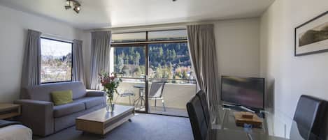 Una sala de estar moderna con un sofá, una mesa de centro y una mesa de vidrio con un televisor. Amplias ventanas ofrecen vistas a árboles y montañas.