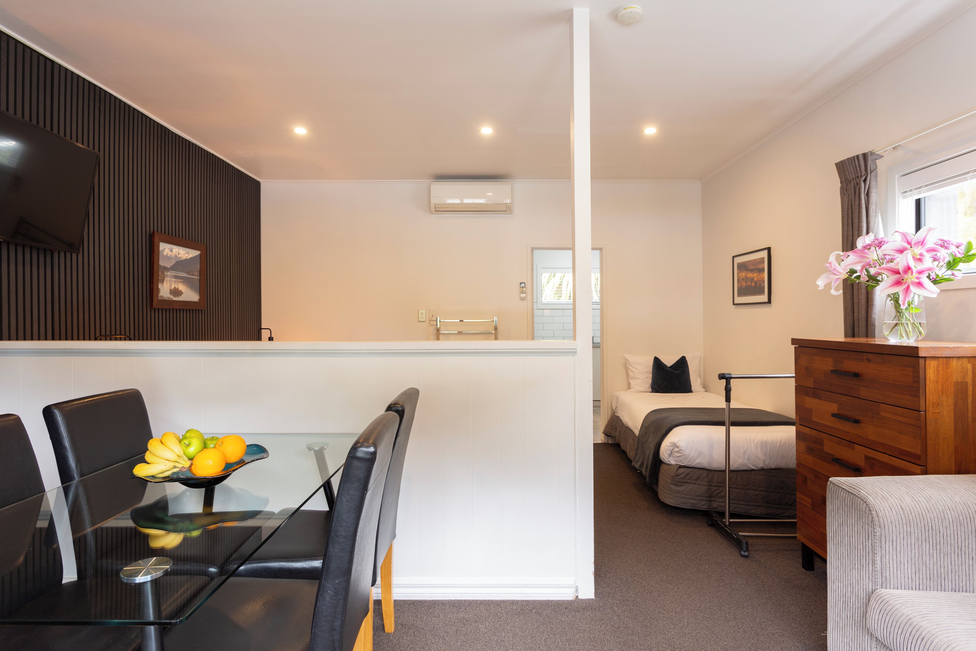 Una habitación de hotel compacta con una mesa de comedor de vidrio, una cama con estructura de metal, un aparador de madera y un pequeño rincón de descanso con una silla.