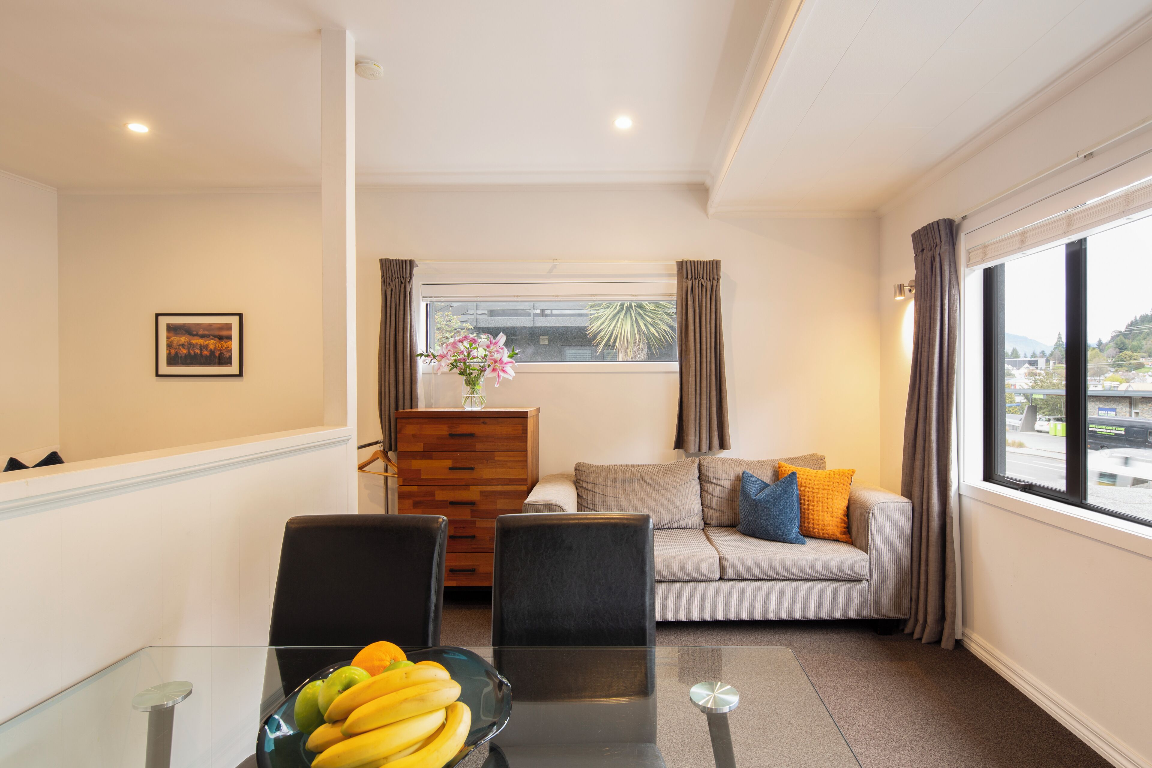 A modern living room with a glass dining table, a sofa with cushions, a wooden cabinet, and a window with curtains.