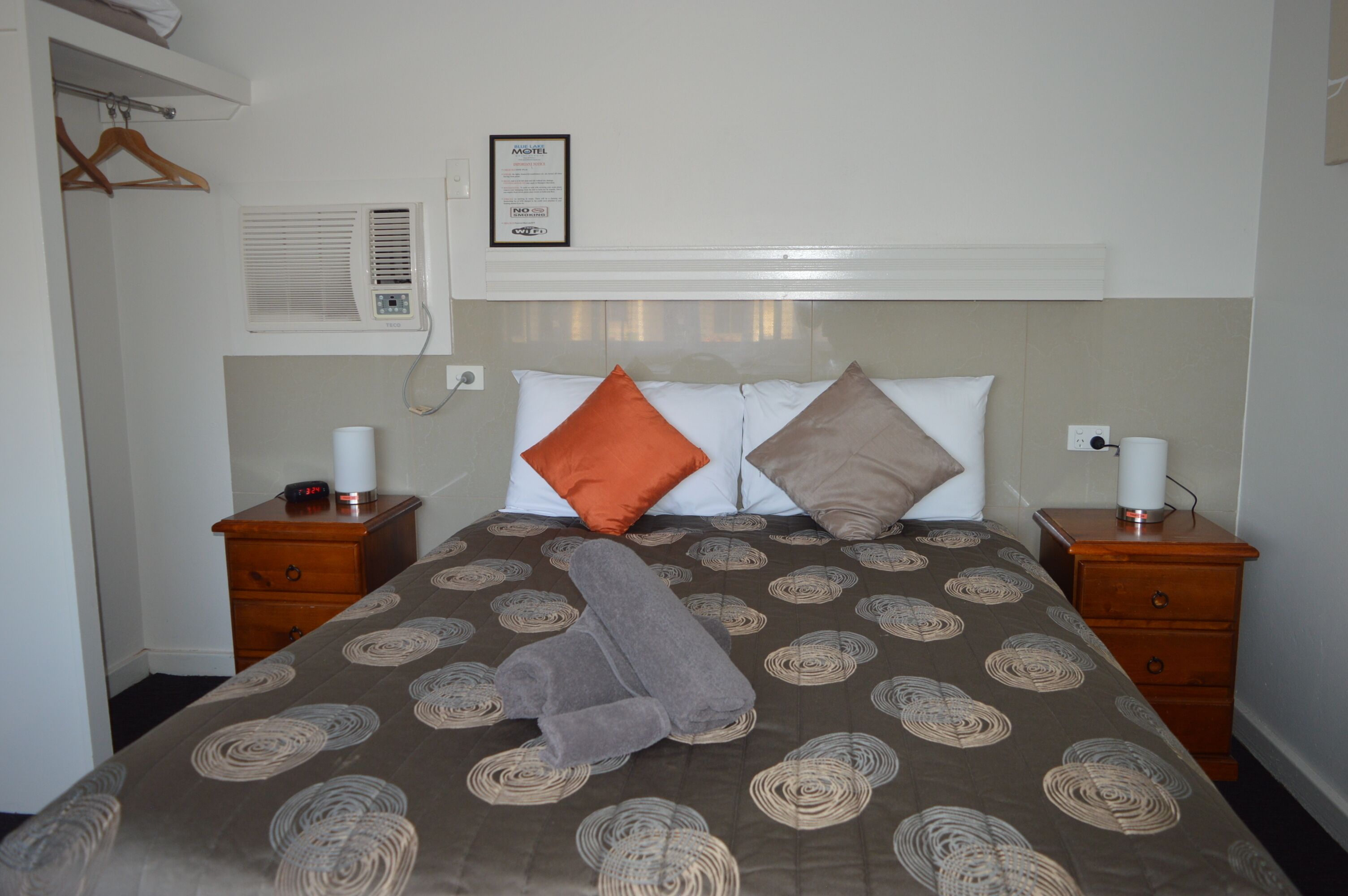 Una habitación de hotel con una cama, dos mesitas de noche, un calefactor, un cartel enmarcado y un perchero de madera.