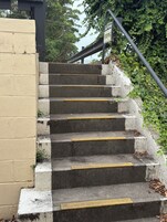 Un conjunto de escalones de concreto con líneas amarillas de seguridad, un letrero de precaución y una barandilla metálica.