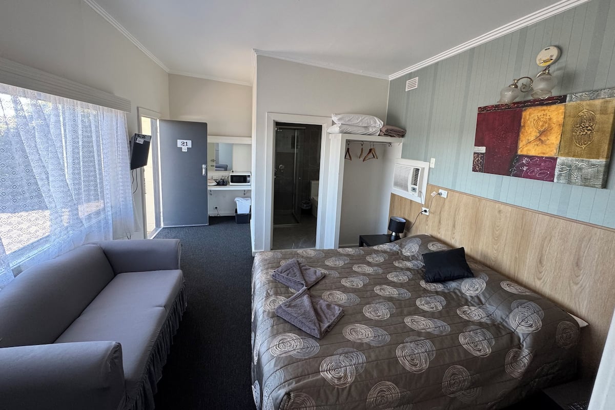 A hotel room with a bed, a sofa, a desk with a chair, a microwave, and a bathroom visible through an open door.