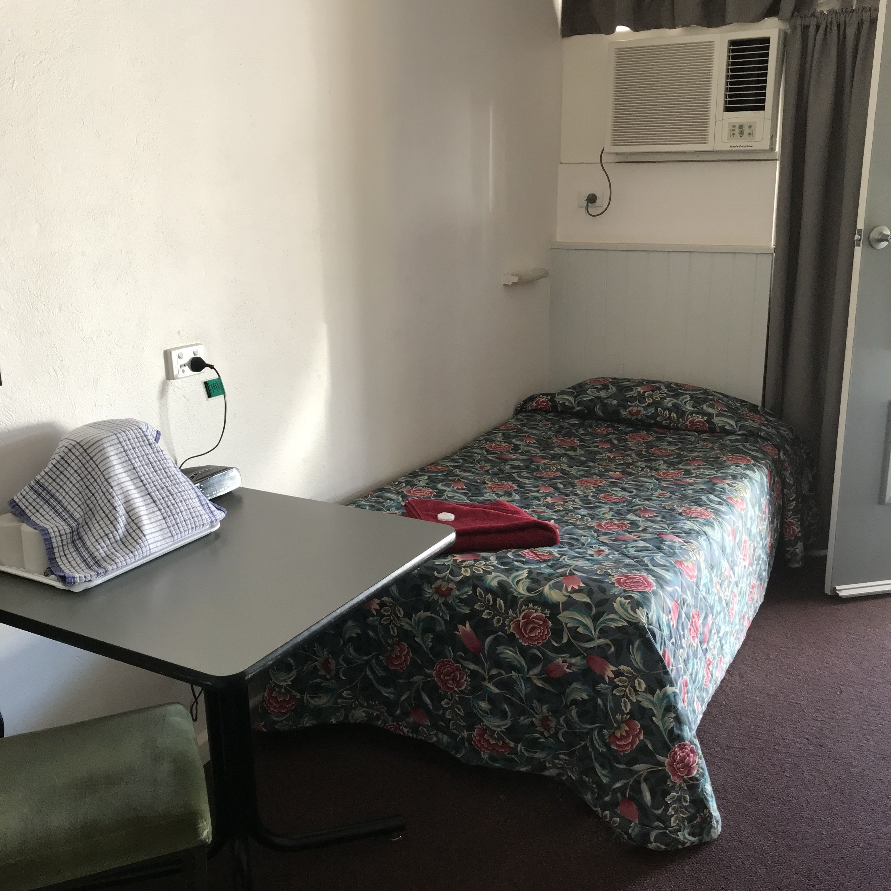 A small, single-bed room with a desk, a pillow, a blanket, and an air conditioning unit.