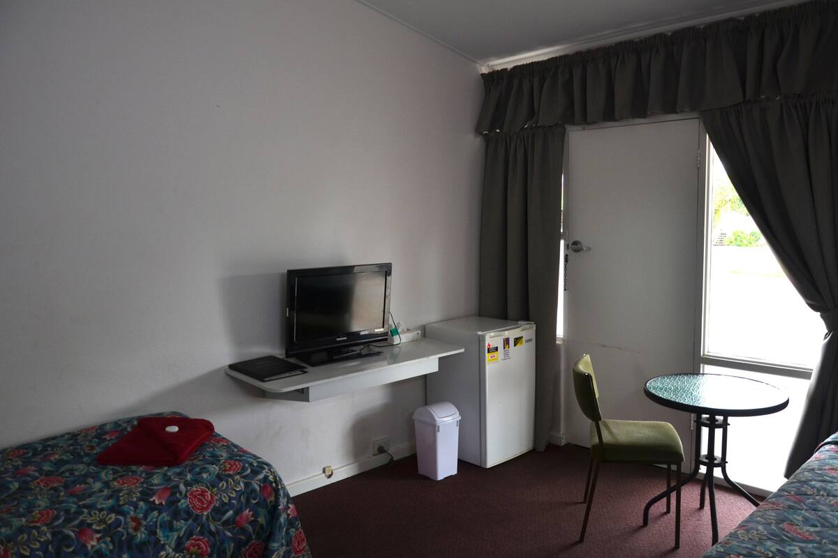 A small hotel room with a bed, a desk with a computer, a refrigerator, a chair, and a window with curtains.