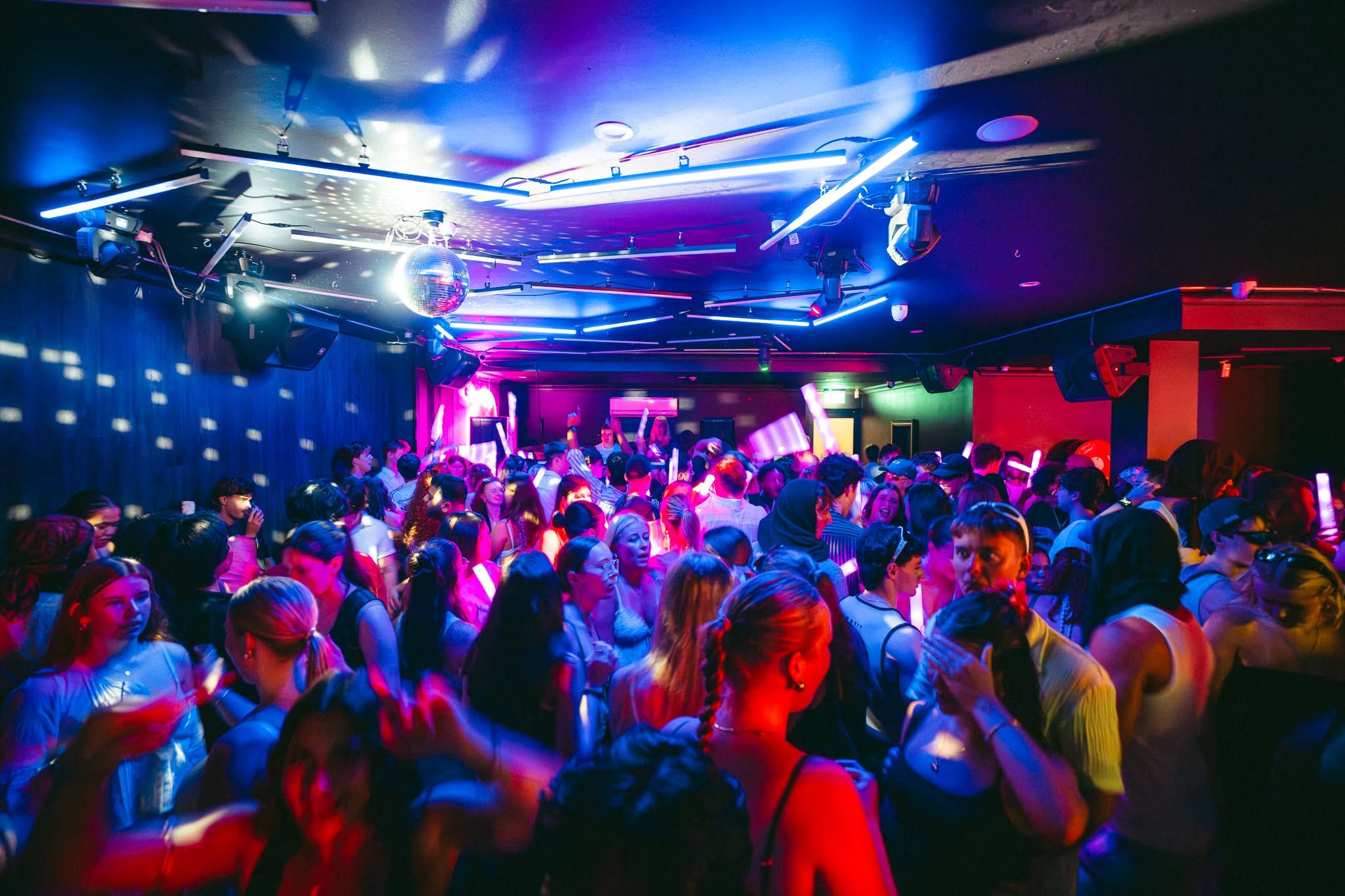 Un espacio interior concurrido donde personas bailan, iluminado por luces de colores.