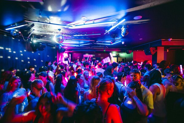 Un espacio interior concurrido donde personas bailan, iluminado por luces de colores.