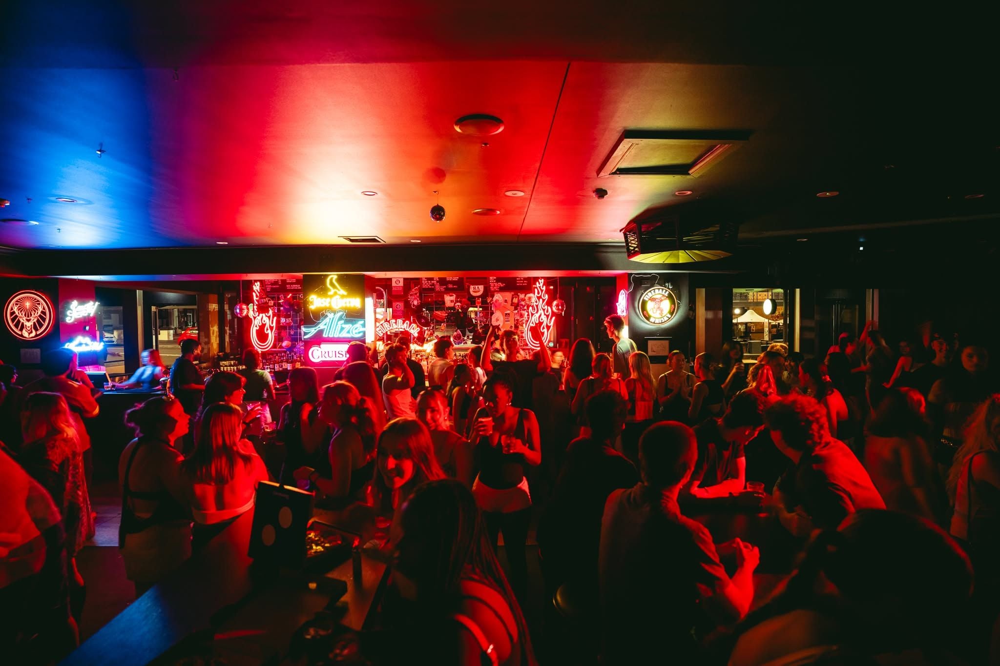 A crowded bar with neon signs and a DJ booth.