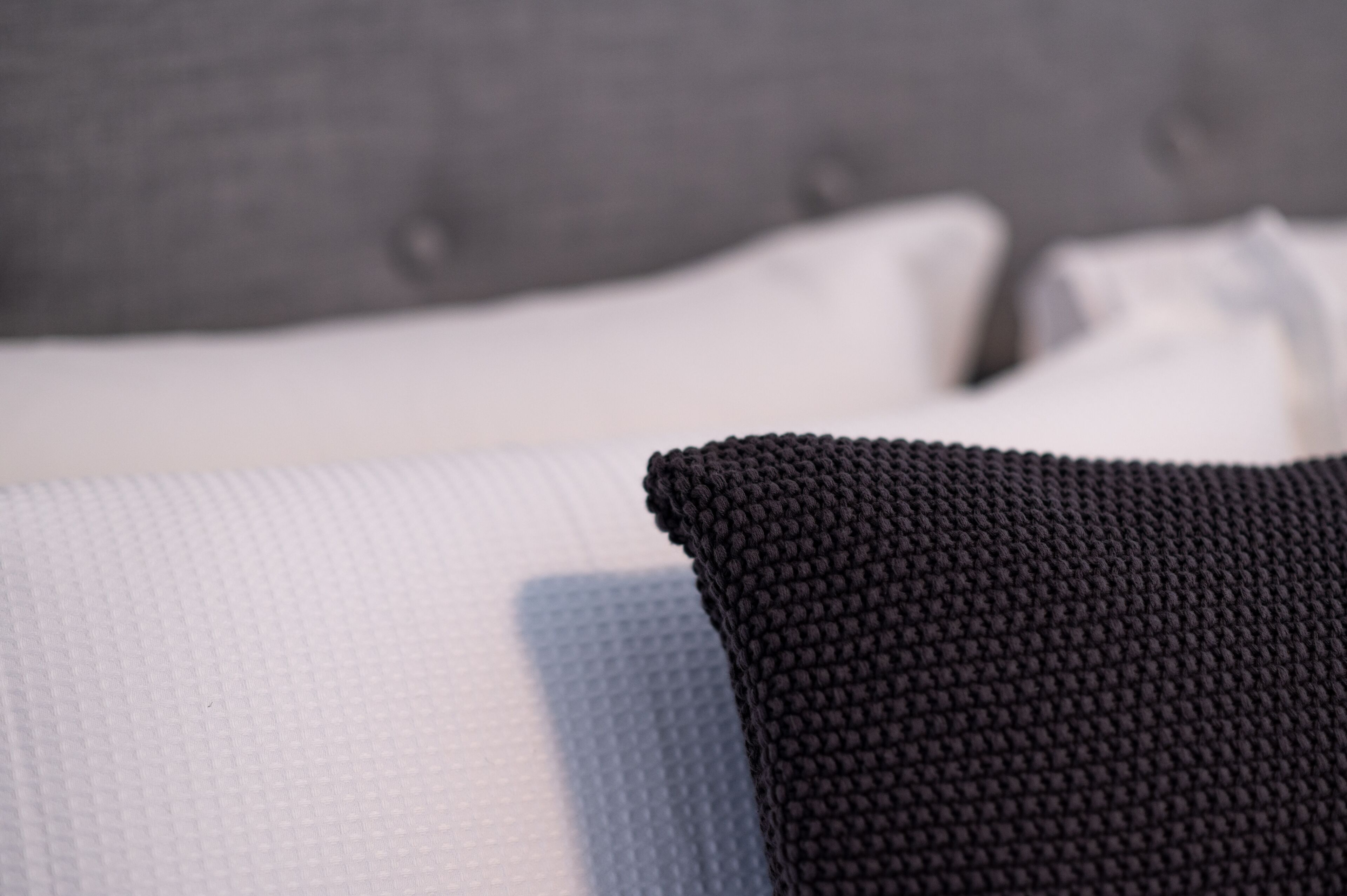 A close-up of a textured white pillow and a black knitted pillow on a bed with a grey headboard.