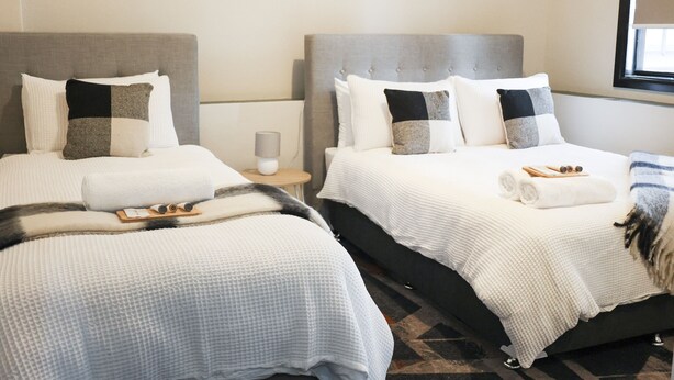 A hotel room with two beds, each with a grey headboard, white bedding, and a small bedside table with a lamp. - Harp Hotel (Wollongong)