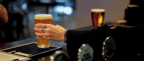 A hand holding a glass of beer at a bar with a beer tap and coaster visible.
