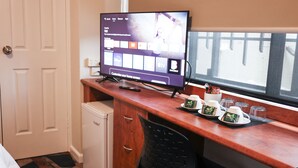 A hotel room with a wooden desk, a television displaying a streaming service interface, a small refrigerator, and a window with a view of a fence. - Harp Hotel (Wollongong)