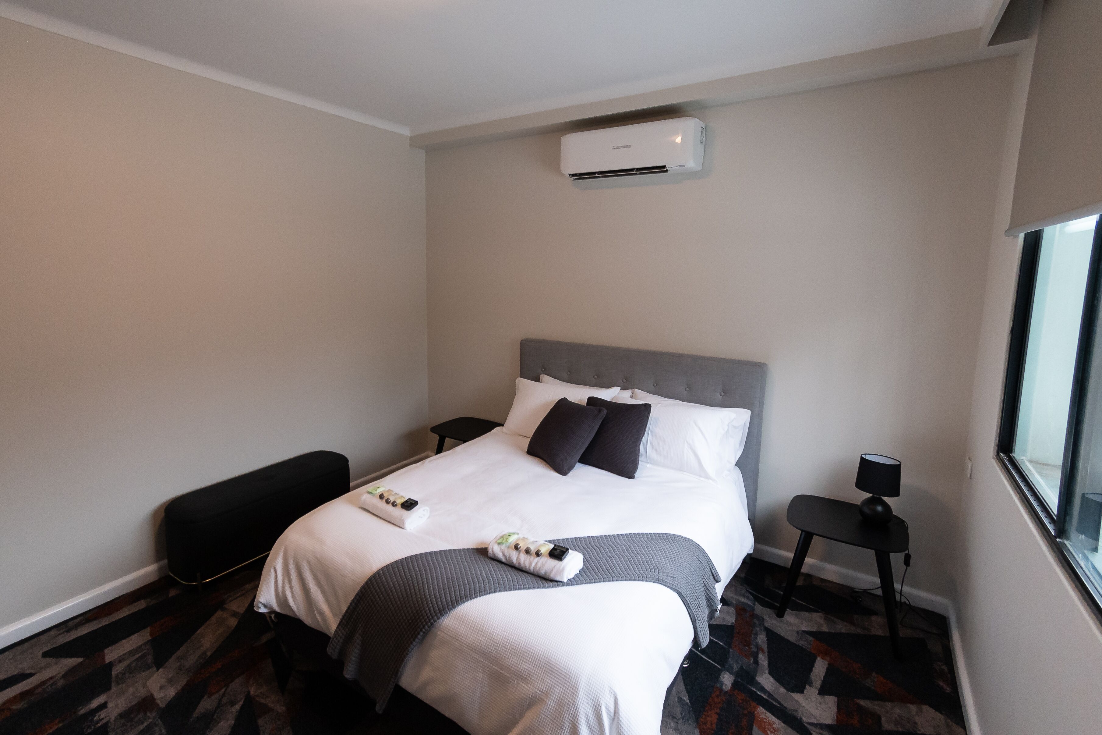 A hotel room with a bed, bedside tables, a bench, an air conditioning unit, and a window.