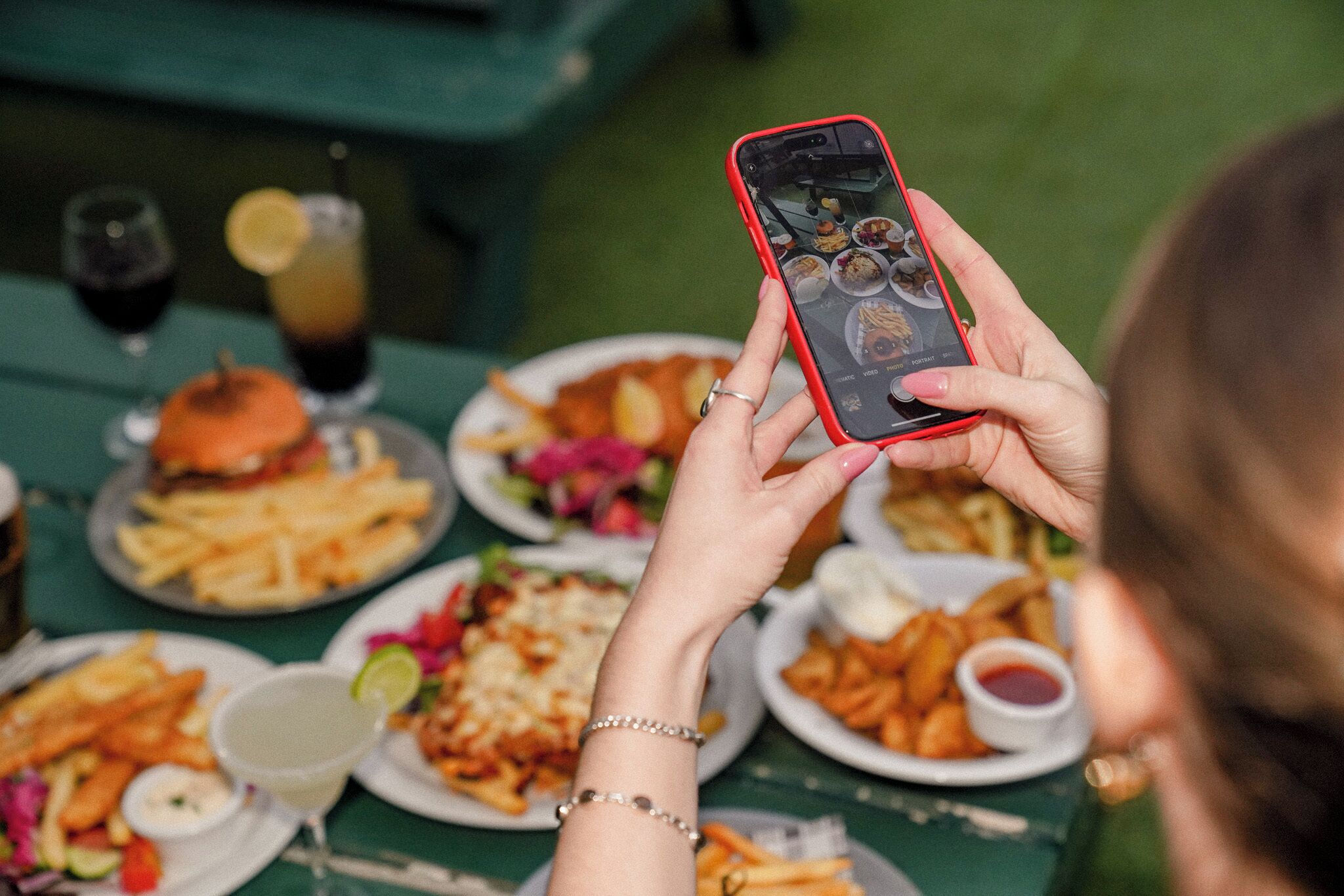Una persona sosteniendo un teléfono inteligente en una mesa con varios platos, incluyendo papas fritas, hamburguesas y bebidas.