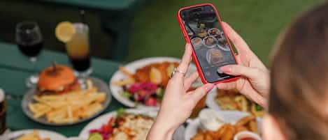 Una persona sosteniendo un teléfono inteligente en una mesa con varios platos, incluyendo papas fritas, hamburguesas y bebidas.