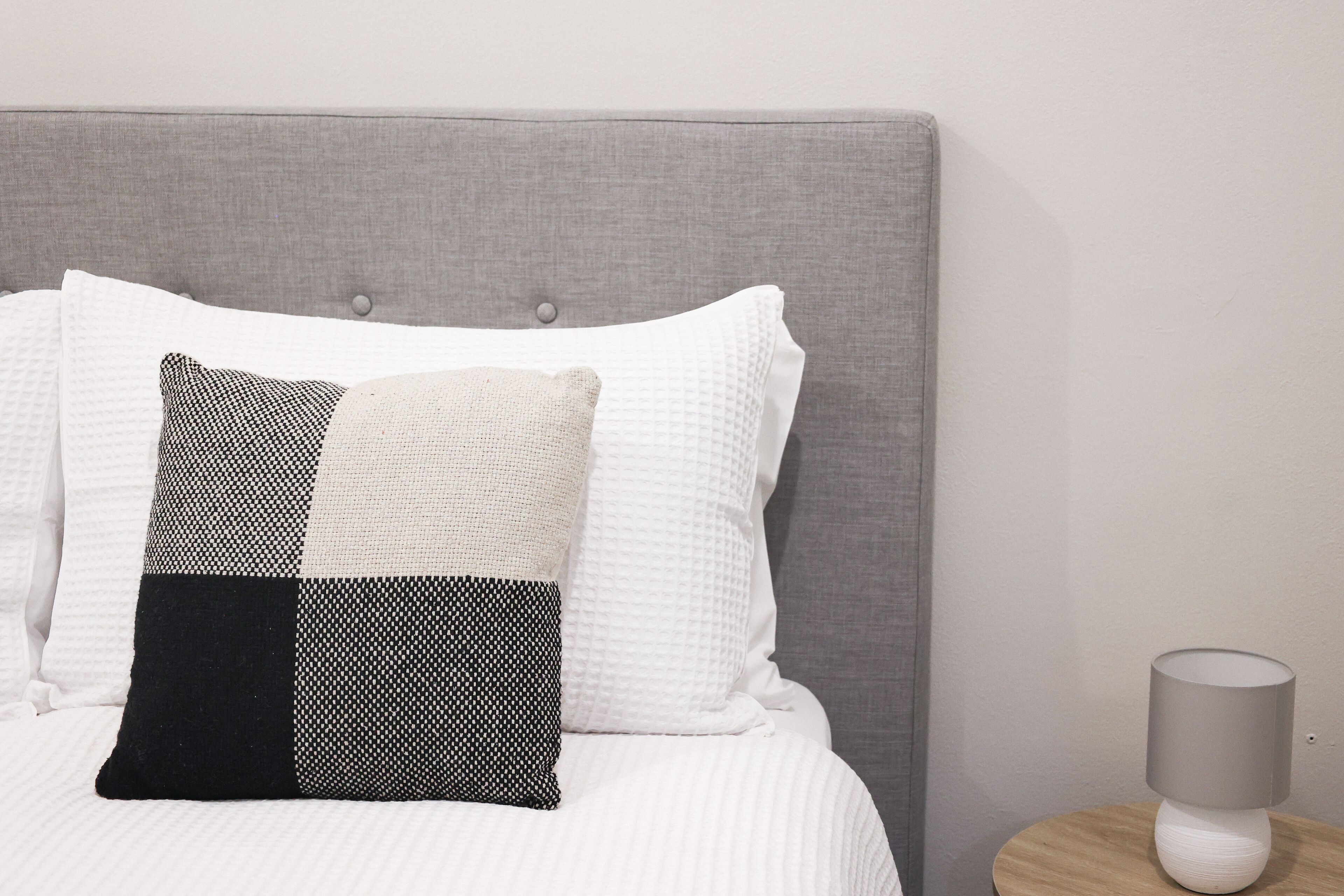Una cama bien hecha con un cabecero tapizado de gris, almohadas blancas y estampadas, y una mesita de noche con una lámpara.