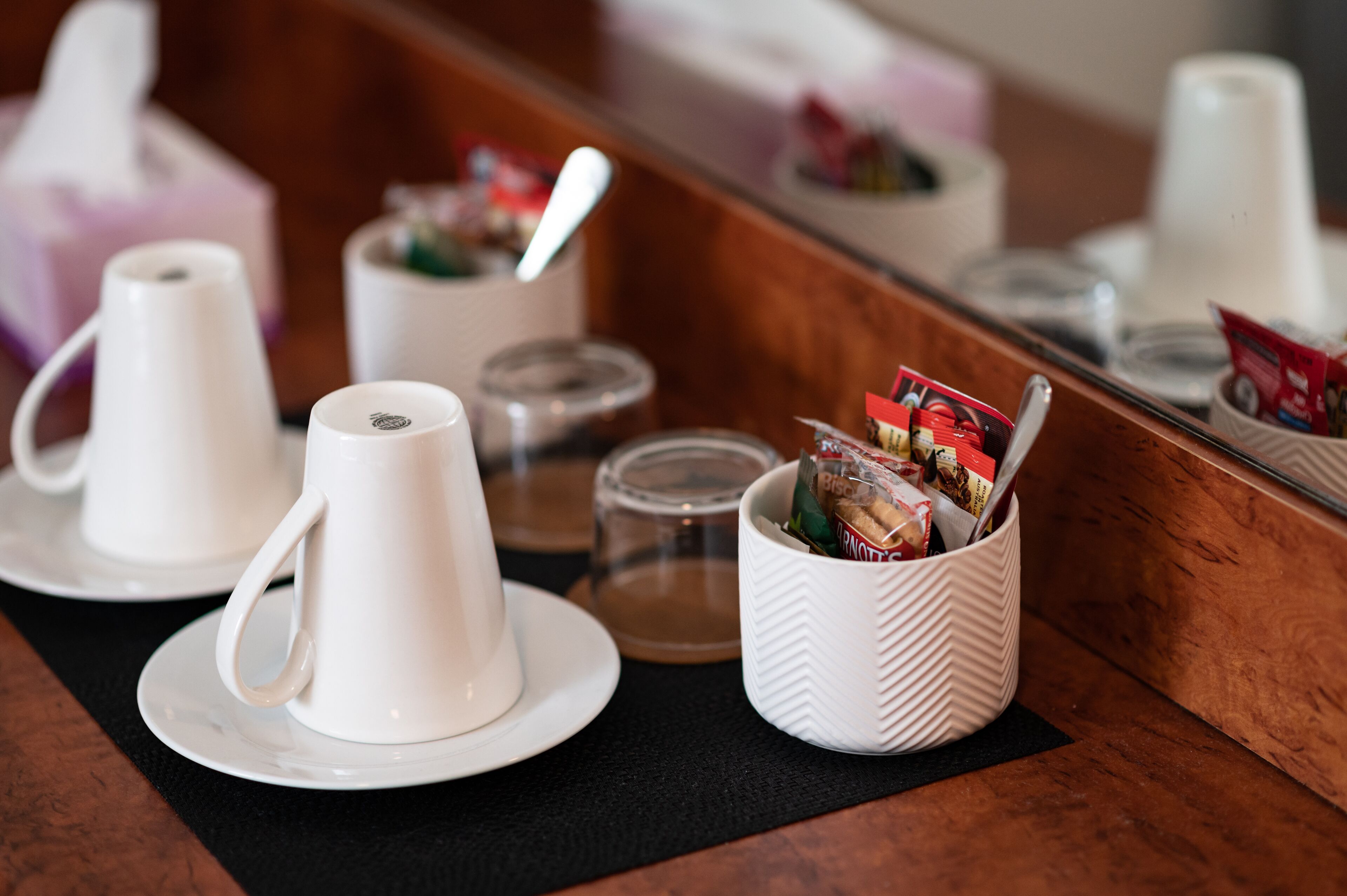 A breakfast setup with white cups and saucers, a glass container with a spoon, and a basket with packaged food items.