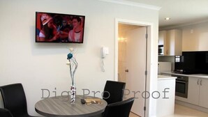 A modern living room with a round table, black chairs, a flat-screen TV mounted on the wall, and a kitchen area visible through an open door. - Auckland Phoenix Palm (Auckland)