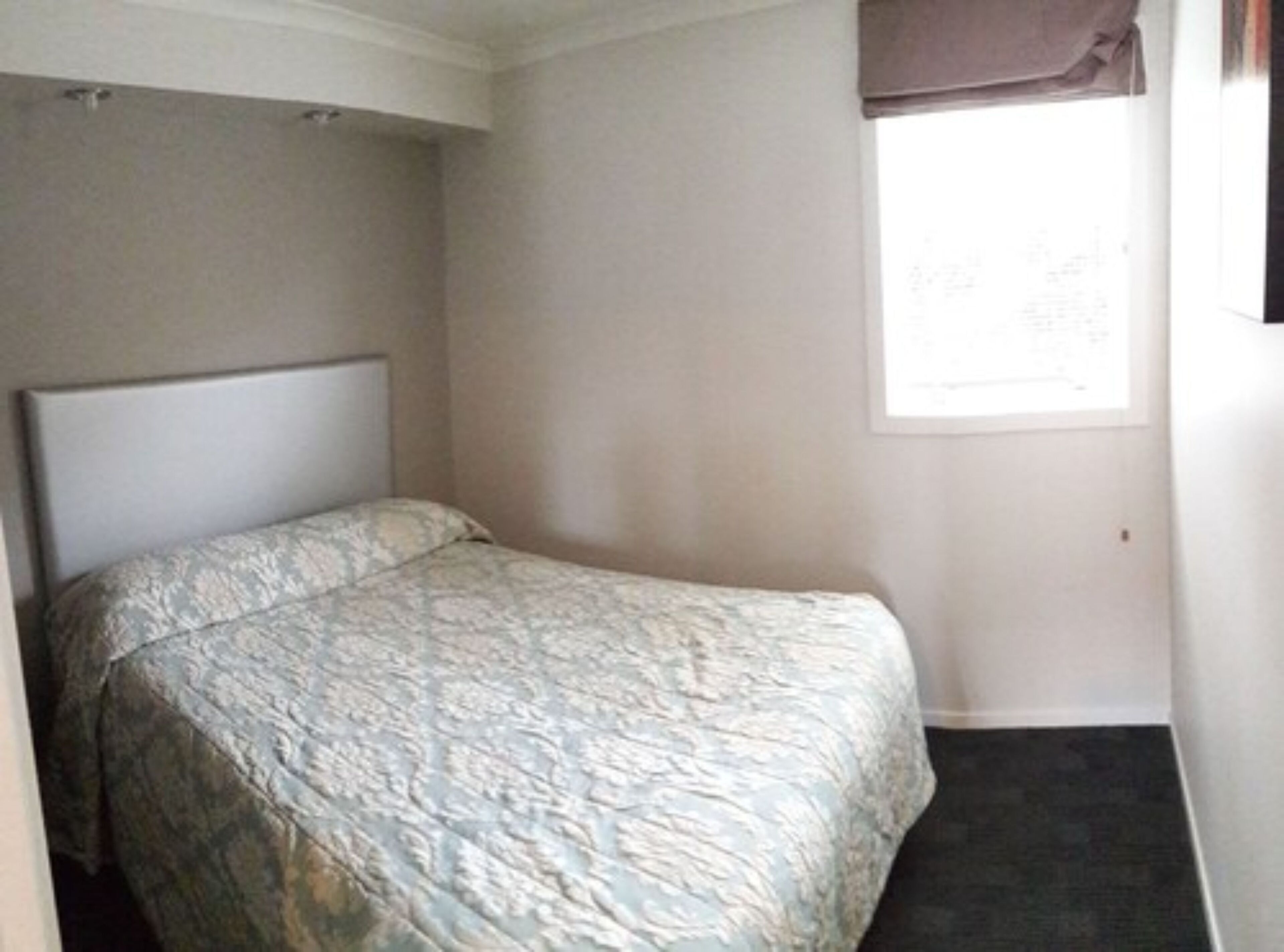 A small, single-bed room with a plain headboard, a window with a brown blind, and a dark carpeted floor.