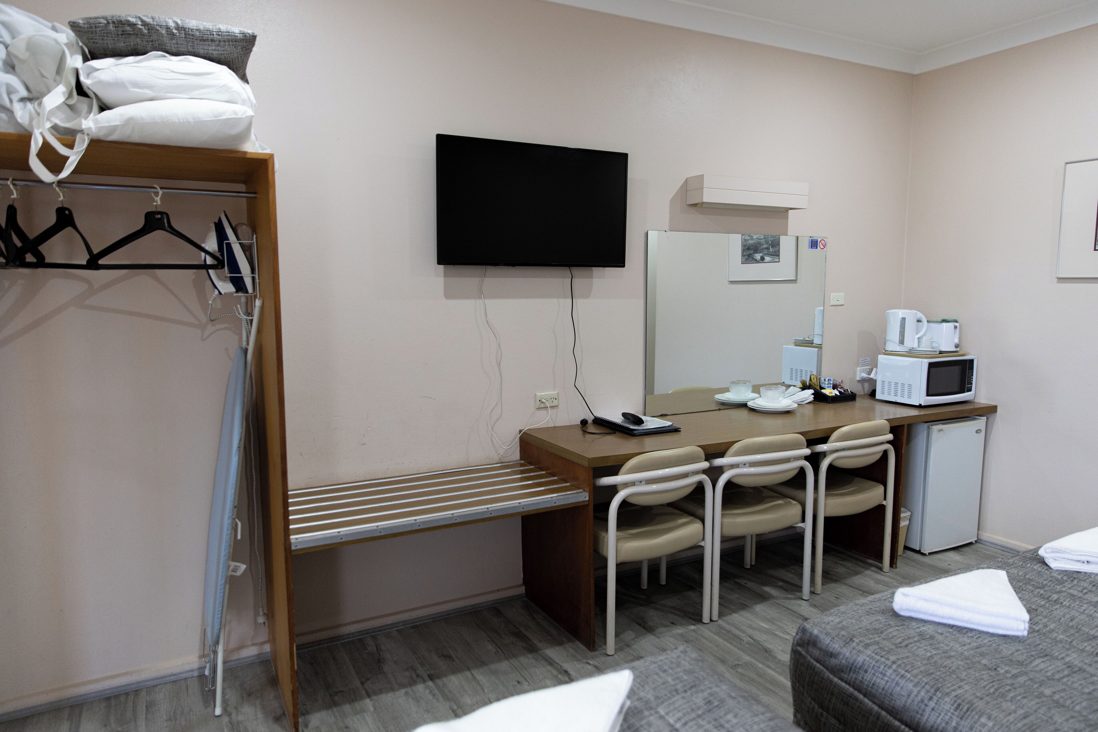 A hotel room with a bed, a desk with a chair, a microwave, a refrigerator, a TV, and a wardrobe with clothes.
