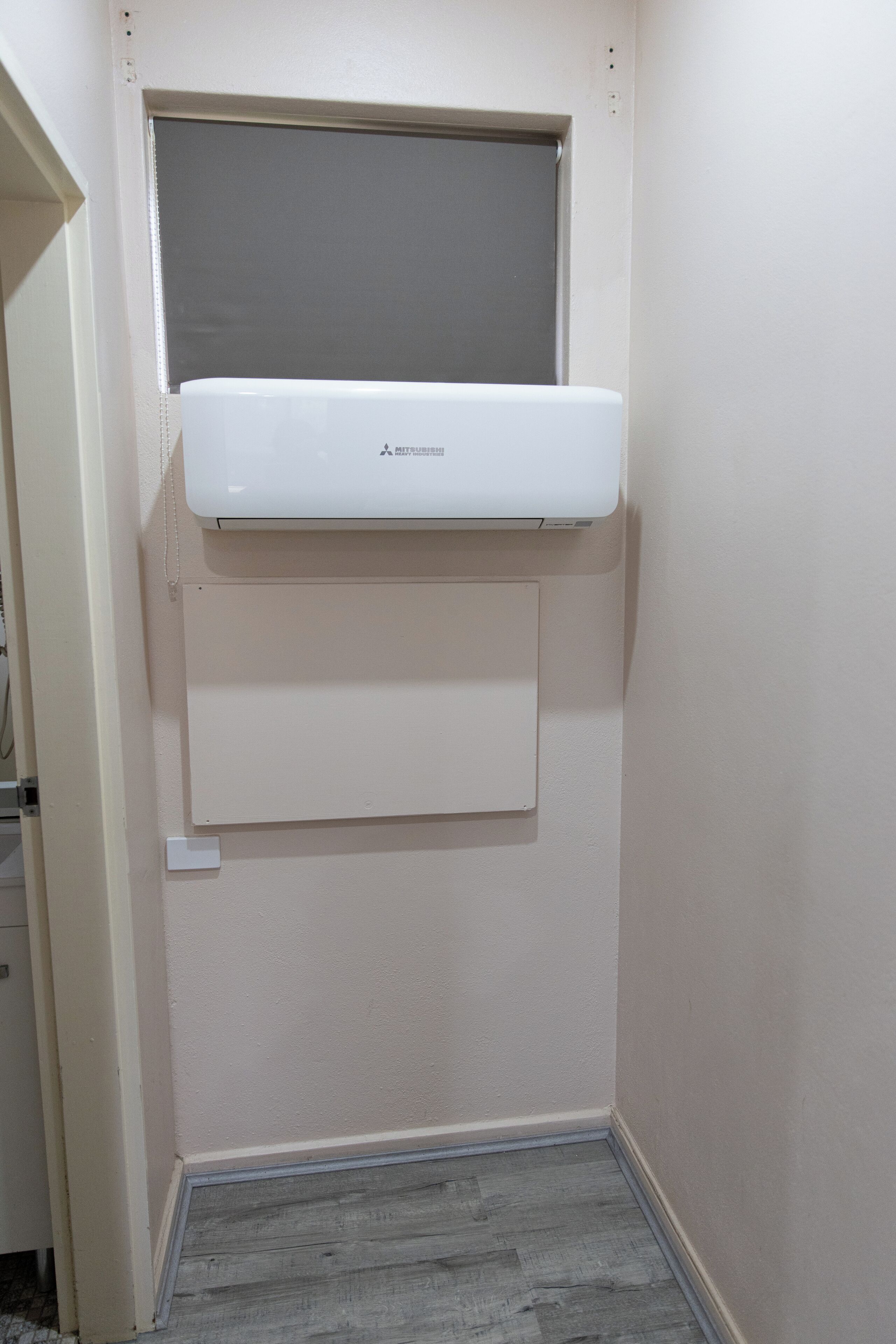 Ein Zimmer mit einer weißen Wand-Klimaanlage, einem kleinen Fenster darüber und einem grauen Holzboden.