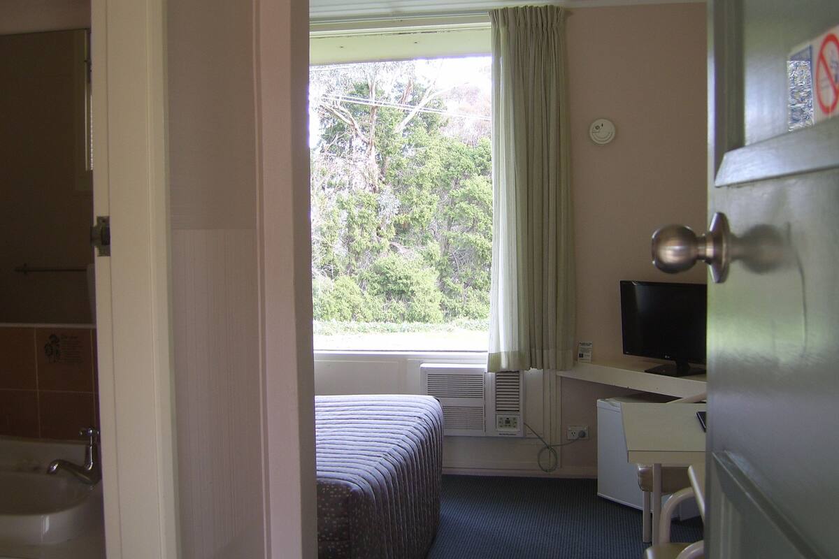 A hotel room with a bed, a desk with a computer, a window with curtains, and a bathroom with a sink and shower.