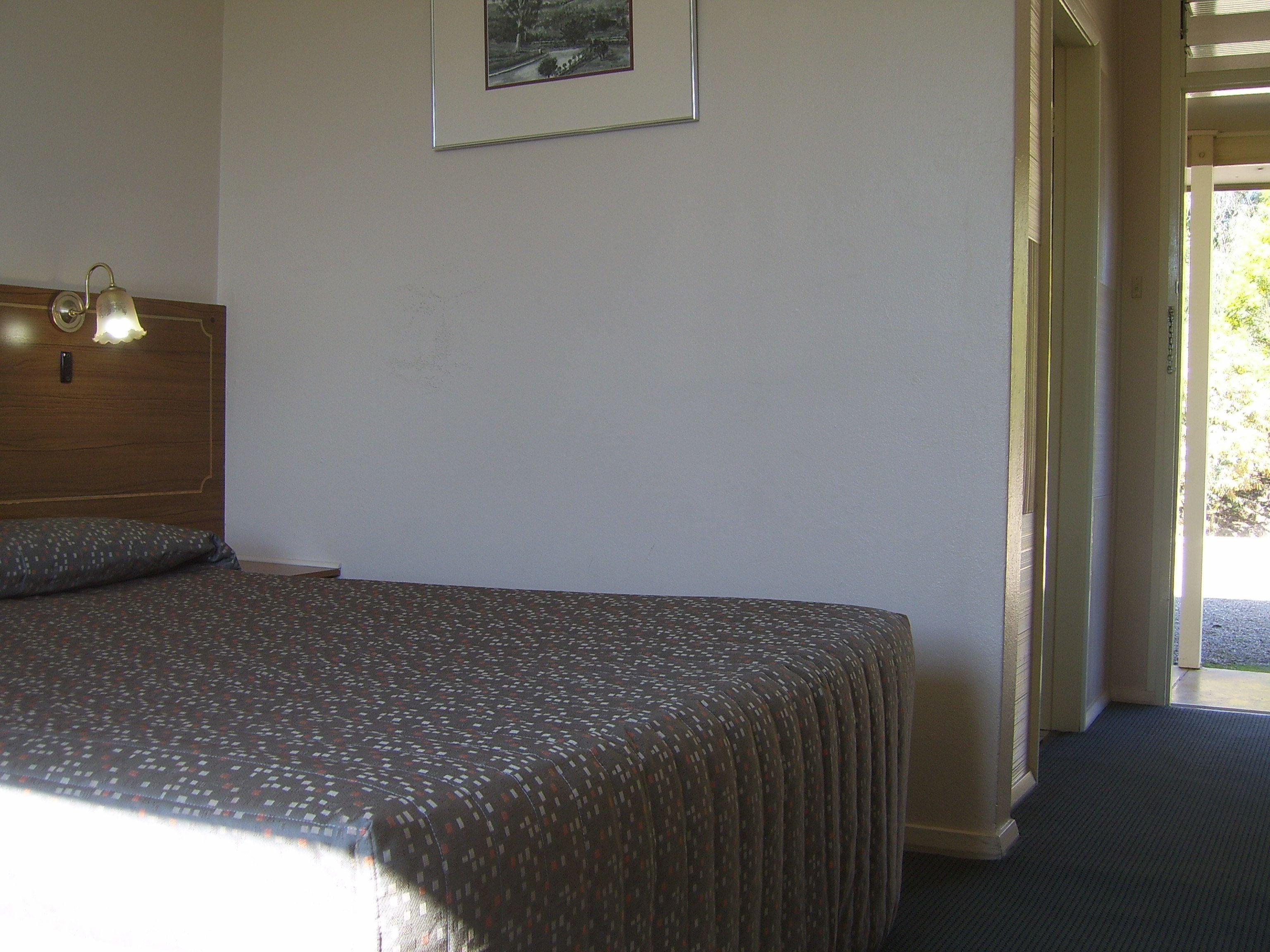 Un letto singolo con una coperta a motivi, un poggiatesta in legno, un quadro incorniciato appeso al muro e un'apertura che dà sull'esterno.