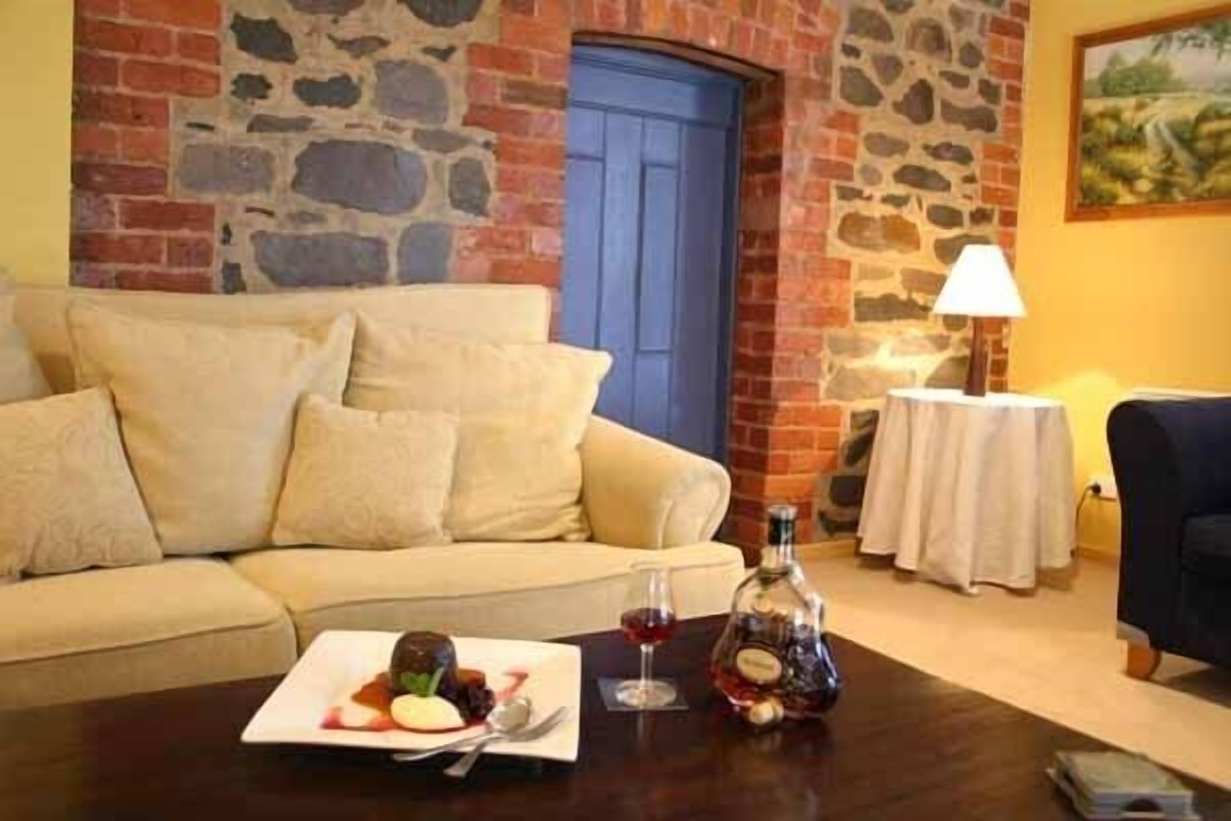 Una habitación acogedora con una pared de piedra, una puerta azul, un sofá color beige, una pequeña mesa con una botella y vasos, y un cuadro en la pared.