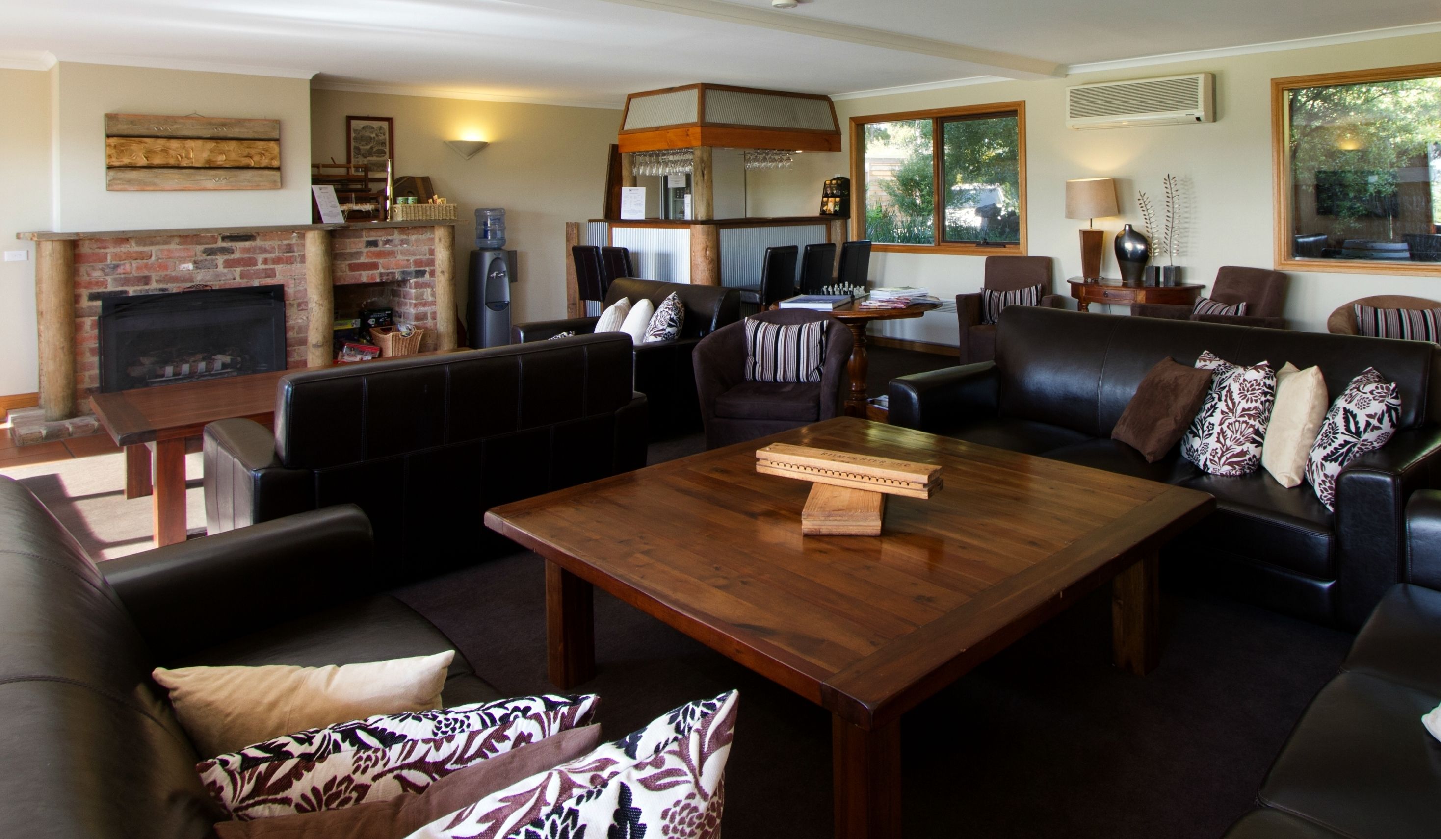 A spacious living room with a fireplace, leather sofas, a wooden coffee table, and a large window offering a view of greenery.