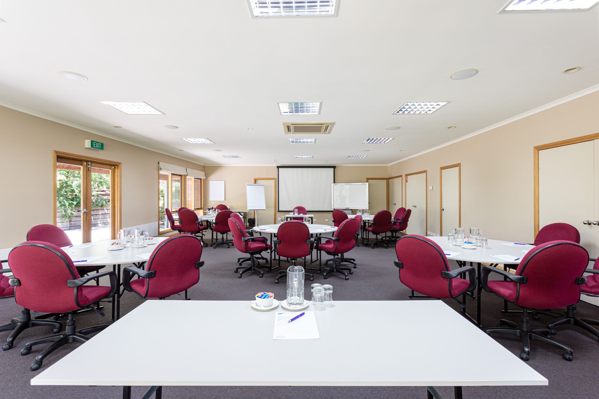 A conference room with multiple tables and red chairs, equipped with glassware and a projector screen.