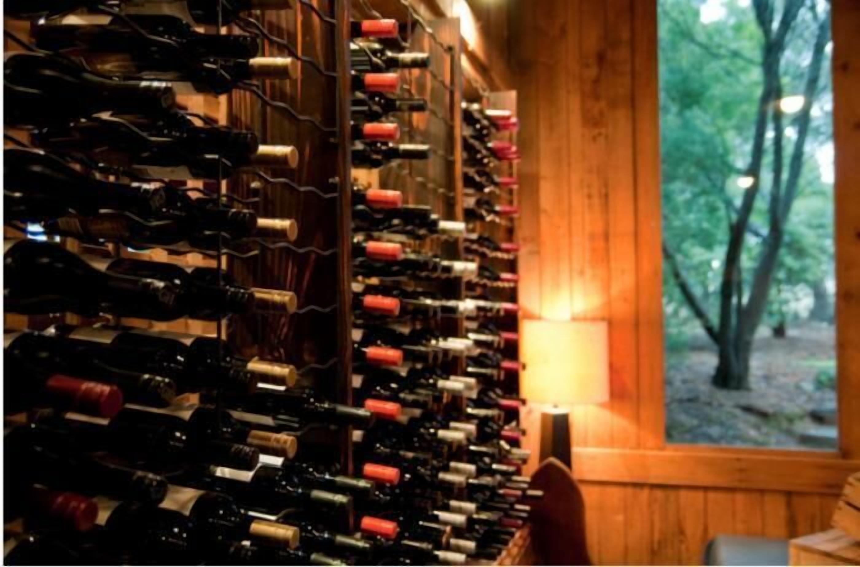 Un celler de vi amb nombroses ampolles organitzades sobre prestatgeries de fusta, interior de fusta i vista d'arbres a través d'una finestra.
