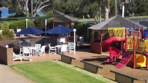 A playground with slides and climbing structures, surrounded by a fenced area with tables and chairs.