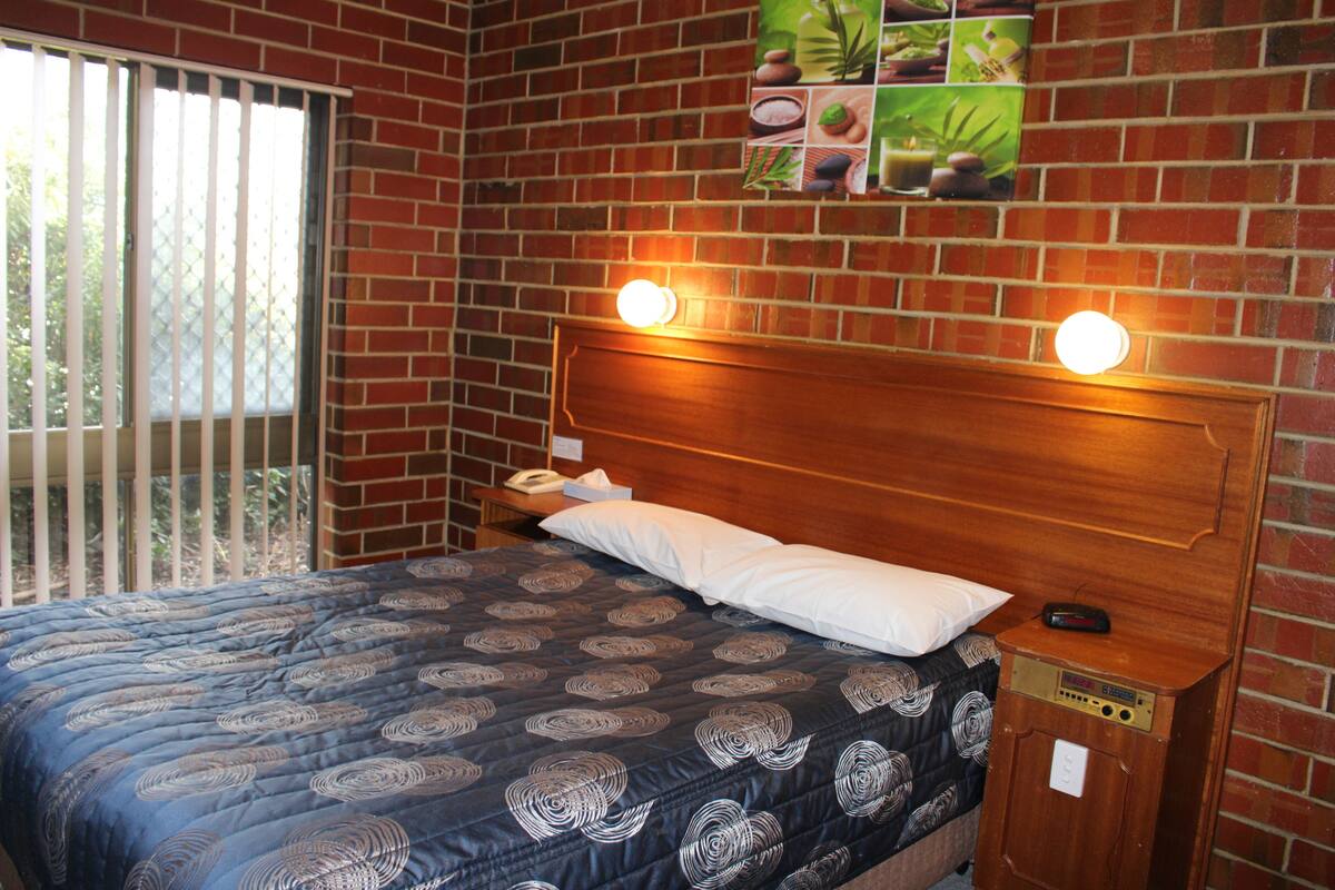 Una habitación con pared de ladrillo, una cama con colcha estampada, dos lámparas de noche y una pequeña mesita de madera con un control remoto.