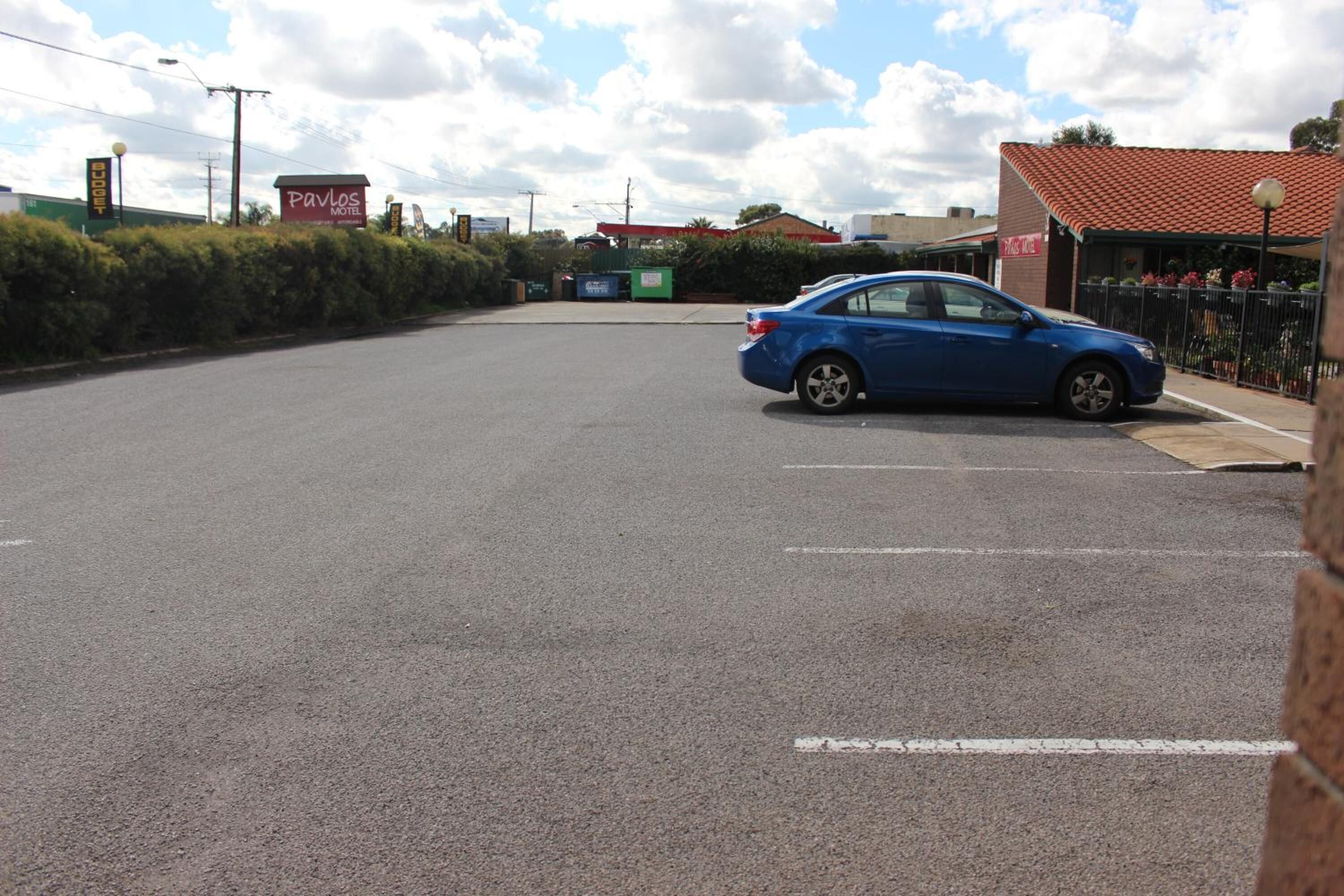 Um estacionamento com um carro azul estacionado perto da entrada de um edifício com telhado vermelho e uma placa que diz 'Pavlos Motel'.