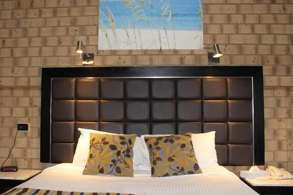 Habitación de hotel con pared de ladrillo, cabecero capitonado, dos almohadas estampadas, mesita de noche con lámpara y un cuadro de una playa.