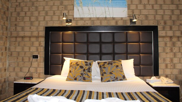 Une chambre d’hôtel avec un mur en briques, un grand lit avec une literie à rayures, deux coussins décoratifs, une tête de lit dotée d’un luminaire et un tableau représentant des herbes de plage.