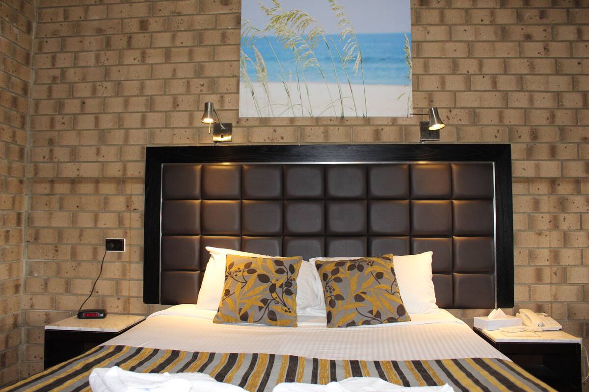 Una habitación de hotel con pared de ladrillo, una cama grande con ropa de cama a rayas, dos almohadas decorativas, un cabecero con una lámpara y un cuadro de hierbas marinas.