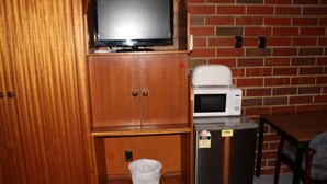 Une petite cuisine équipée d’un four à micro-ondes, d’un petit réfrigérateur, d’un téléviseur posé sur un meuble en bois et d’un récipient en plastique sur le plan de travail.