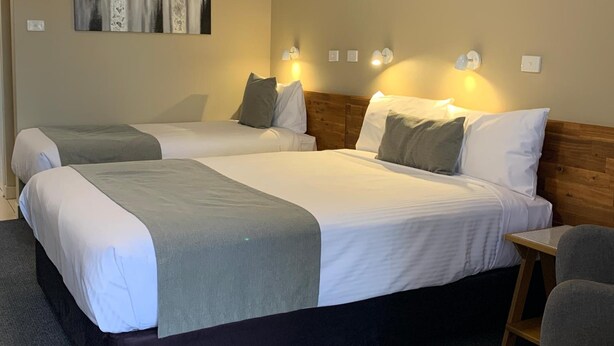 A hotel room with two beds, a wooden headboard, a small wooden table, and wall-mounted lights. - In Town Motor Inn (Taree)