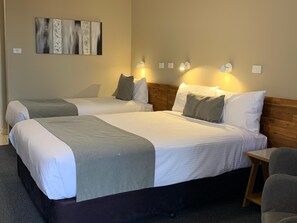 A hotel room with two beds, a wooden headboard, a small wooden table, and wall-mounted lights. - In Town Motor Inn (Taree)